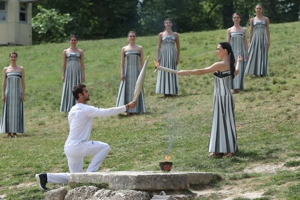 扮演最高女祭司的演員瑪麗·米娜（前右）點(diǎn)燃第一棒火炬手——希臘運(yùn)動(dòng)員、東京奧運(yùn)會(huì)男子賽艇項(xiàng)目金牌得主斯特凡諾斯·恩圖斯科斯手中的火炬。新華社記者 趙丁喆 攝